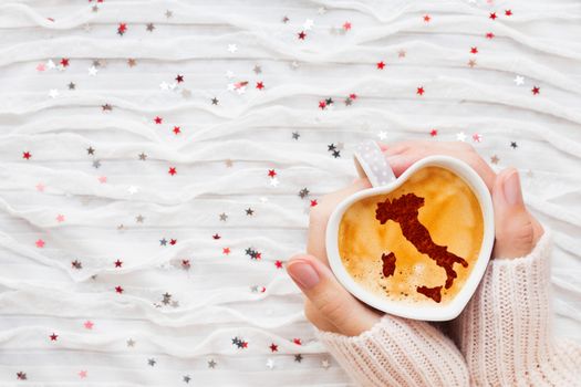 Woman holds a cup of hot coffee with cinnamon silhouette of Italy. Winter wanderlust and Valentine's Day fabric background with sparkling silver and red confetti.
