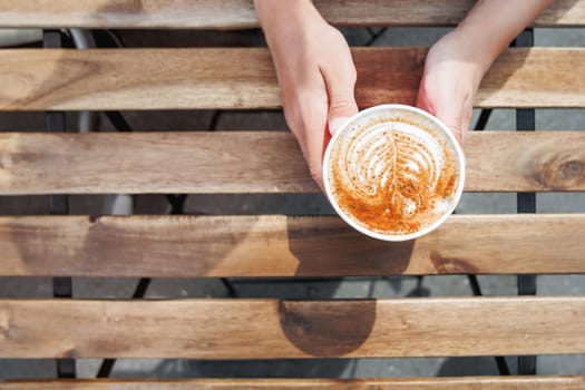 Woman holding a paper cup with coffee. Coffee to go. Tasty hot beverage on wooden table in sunny day. Outdoors meal. Flat lay, top view.