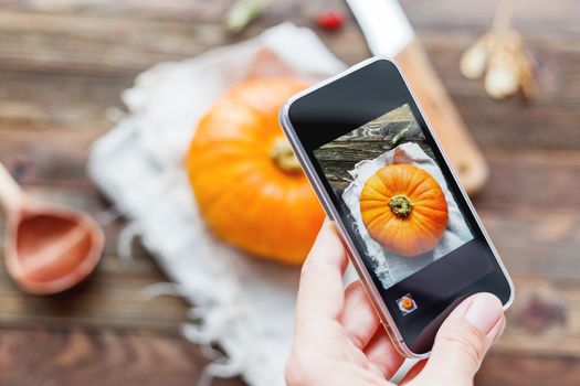 Woman shooting fresh orange pumpkin on wooden table using smartphone. Homespun napkin and rustic cutlery on background.