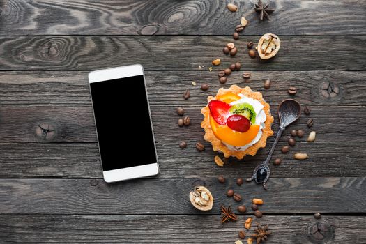Wooden rustic background with fruit tart and spices - peanuts, anise stars, coffee beans, walnuts. Tasty dessert with strawberry, kiwi, orange, peach and whipped cream. Smartphone, good for mock up.