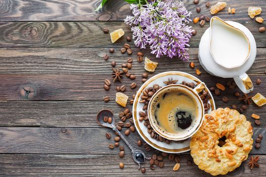 Rustic wooden background with cup of coffee, milk, peanut tart, sugar ginger and lilac flowers. White vintage dinnerware and spoon. Breakfast at summer morning. Top view, place for text.