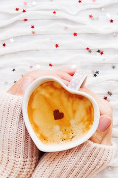 Woman holds a cup of hot coffee with cinnamon heart. Winter and Valentine's Day fabric background with sparkling silver and red confetti.