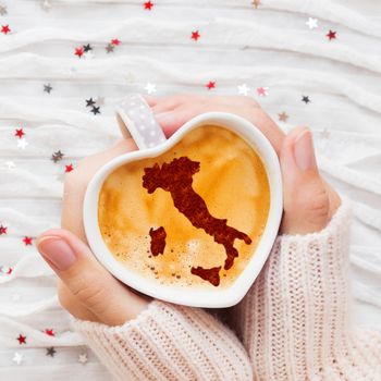 Woman holds a cup of hot coffee with cinnamon silhouette of Italy. Winter wanderlust and Valentine's Day fabric background with sparkling silver and red confetti.