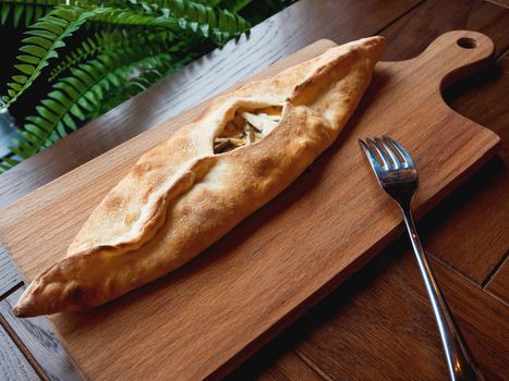 Baked bun in the shape of a boat with mozzarella cheese, cheddar cheese and oyster mushrooms. Pastry on wooden chopping board.