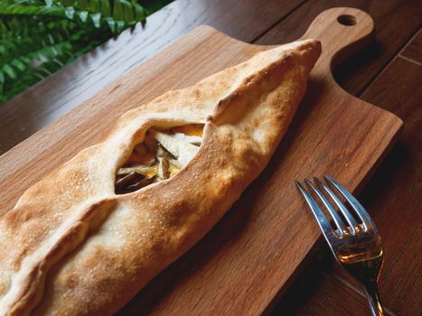 Baked bun in the shape of a boat with mozzarella cheese, cheddar cheese and oyster mushrooms. Pastry on wooden chopping board.