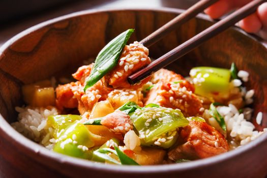 Asian cuisine - rice in sauce with stir fried vegetables, pineapple and salmon. Wooden bowl with chopsticks. Close up.