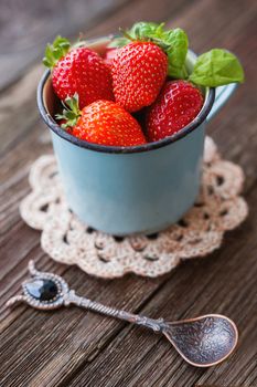 Fresh juicy strawberries in old rusty mug. Rustic wooden background with croshet napkin and vintage spoon.