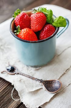 Fresh juicy strawberries in old rusty mug. Rustic wooden background with homespun napkin and vintage spoon.