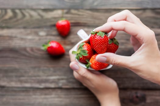 Woman holds a cup full of strawberries in hands. Mug shaped as a heart, symbol of love. Fresh juicy strawberry on rustic wooden background. Top view, place for text.