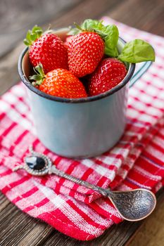 Fresh juicy strawberries in old rusty mug. Rustic wooden background with plaid napkin and vintage spoon.