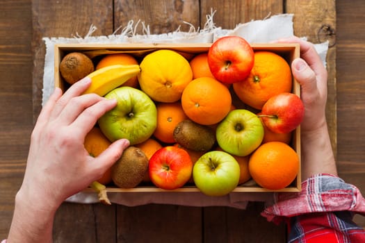 Man in tartan plaid shirt holds a box full of fresh fruits. Fruit harvest - apples, oranges, lemon, kiwi, banana. Rustic wooden table.