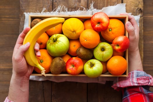 Man in tartan plaid shirt holds a box full of fresh fruits and a banana. Fruit harvest - apples, oranges, lemon, kiwi, banana. Rustic wooden table.