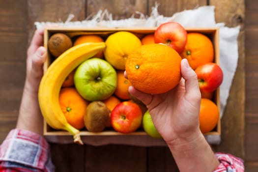 Man in tartan plaid shirt holds a box full of fresh fruits and a big juicy orange. Fruit harvest - apples, oranges, lemon, kiwi, banana. Rustic wooden table.