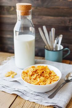 Tasty corn flakes in bowl with bottle of milk. Rustic wooden background with homespun napkin. Healthy crispy breakfast snack.