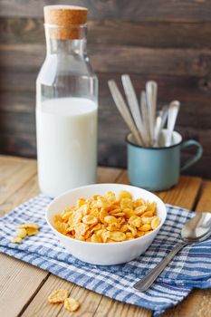 Tasty corn flakes in white bowl with bottle of milk. Rustic wooden background with plaid blue napkin. Healthy crispy breakfast snack.