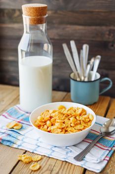 Tasty corn flakes in white bowl with bottle of milk. Rustic wooden background with plaid napkin. Healthy crispy breakfast snack.