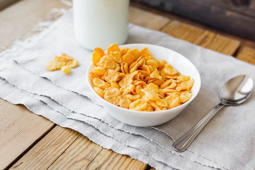 Tasty corn flakes in bowl with bottle of milk. Rustic wooden background with homespun napkin. Healthy crispy breakfast snack.