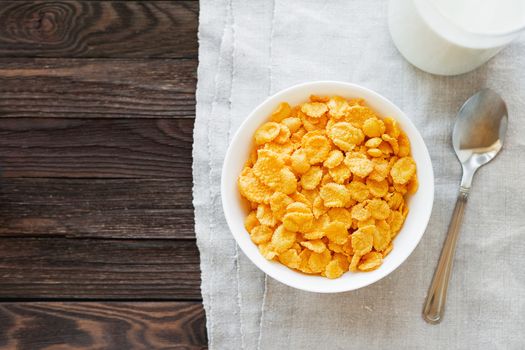 Tasty corn flakes in bowl with bottle of milk. Rustic wooden background with homespun napkin. Healthy crispy breakfast snack. Place for text. Top view, flat lay.