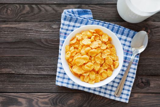 Tasty corn flakes in bowl with bottle of milk. Rustic wooden background with plaid tartan napkin. Healthy crispy breakfast snack. Place for text. Top view, flat lay.