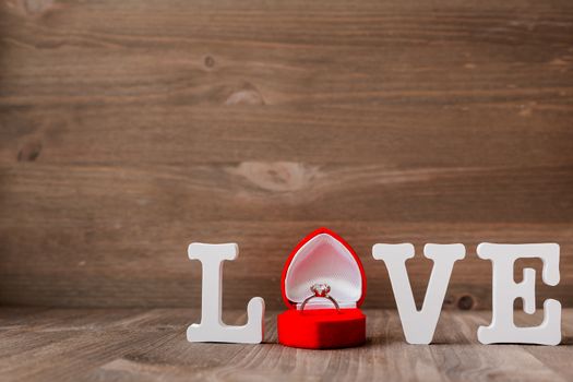 Word LOVE and diamond engagement ring in red gift box on wooden background. Symbol of love and marriage. Good as Valentine's Day card with place for text.