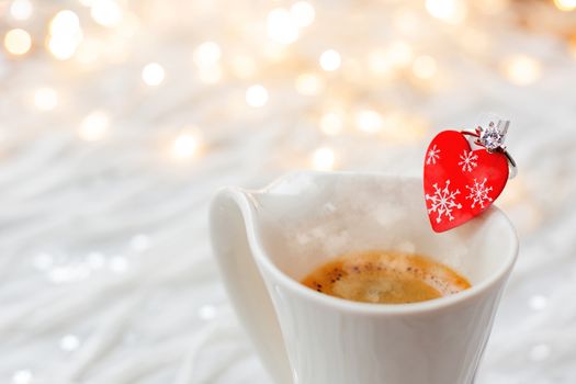 White cup of hot coffee with decorative heart and engagement diamond ring, symbol of love and marriage. Valentine's day background with lights and decorations.