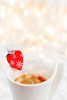 White cup of hot coffee with decorative heart and engagement diamond ring, symbol of love and marriage. Valentine's day background with lights and decorations.