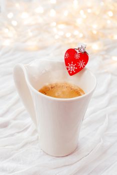 White cup of hot coffee with decorative heart and engagement diamond ring, symbol of love and marriage. Valentine's day background with lights and decorations.