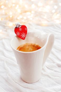 White cup of hot coffee with decorative heart and engagement diamond ring, symbol of love and marriage. Valentine's day background with lights and decorations.