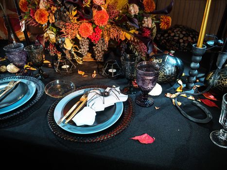 Table served in Halloween style. Bouquet with red flowers, decorative spiders, pumpkins with lettering and gothic vine glasses on black tablecloth. Holiday decorations. Place for text.