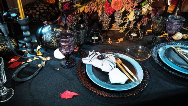 Table served in Halloween style. Bouquet with red flowers, decorative spiders, pumpkins with lettering and gothic vine glasses on black tablecloth. Holiday decorations. Place for text.