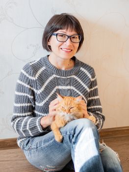 Cute ginger cat dozing on woman knees. Smiling woman in torn jeans stroking her fluffy pet. Cozy home.