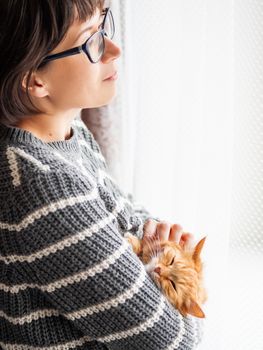 Cute ginger cat dozing on woman arms. Smiling woman in grey knitted sweater stroking her fluffy pet. Cozy home.