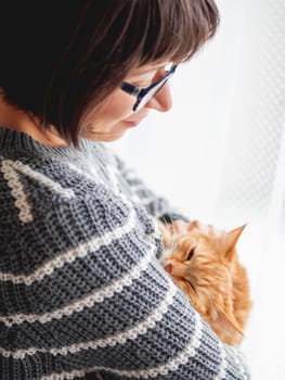 Cute ginger cat dozing on woman arms. Smiling woman in grey knitted sweater stroking her fluffy pet. Cozy home.