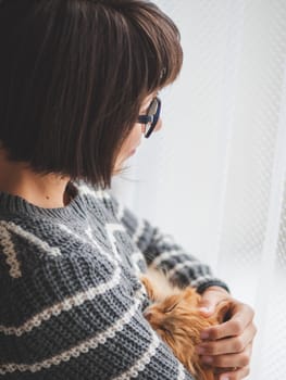 Cute ginger cat dozing on woman arms. Smiling woman in grey knitted sweater stroking her fluffy pet. Cozy home.