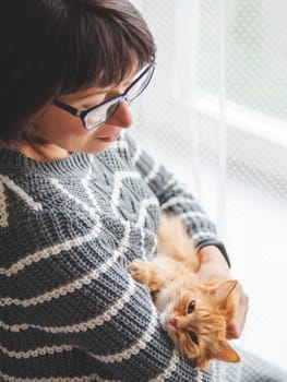 Cute ginger cat dozing on woman arms. Smiling woman in grey knitted sweater holding her fluffy pet. Cozy home.