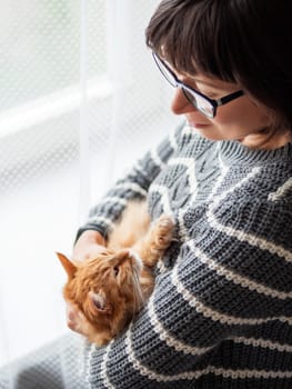Cute ginger cat dozing on woman arms. Smiling woman in grey knitted sweater holding her fluffy pet. Cozy home.