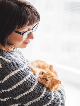 Cute ginger cat dozing on woman arms. Smiling woman in grey knitted sweater stroking her fluffy pet. Cozy home.