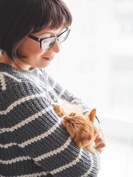 Cute ginger cat dozing on woman hands. Smiling woman in grey knitted sweater stroking her fluffy pet. Cozy home.