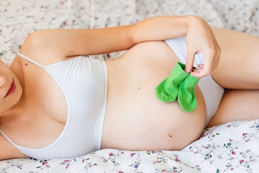 Pregnant woman in white underwear lying in bed with small baby's socks. Young woman expecting a baby.