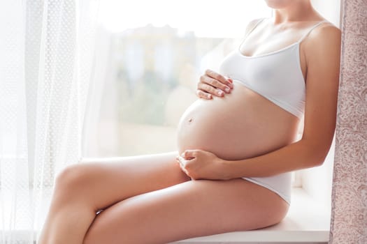 Pregnant woman in white underwear sitting on window sill. Young woman expecting a baby. Cozy happy background in sunny morning.