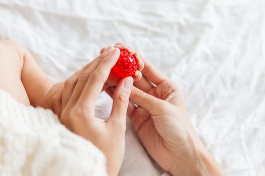 Mother holds newborn baby's feet. Tiny fingers and red massage ball in woman's hand. Cozy morning at home.