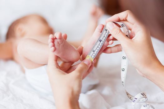 Mother holds newborn baby's bare feet and measures the length of foot. Cozy morning at home.