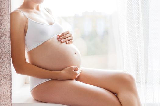 Pregnant woman in white underwear sitting on window sill. Young woman expecting a baby. Cozy happy background in sunny morning.