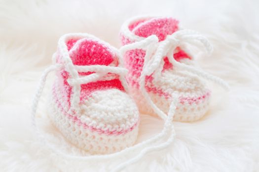Little baby shoes. Hand knitted first sneakers for newborn girl. Crochet handmade pink bootees on fluffy white background.