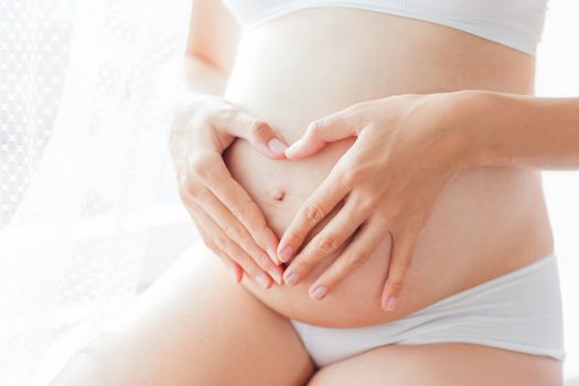 Pregnant woman in white underwear. Young woman expecting a baby. Future mother makes heart gesture - symbol of love.