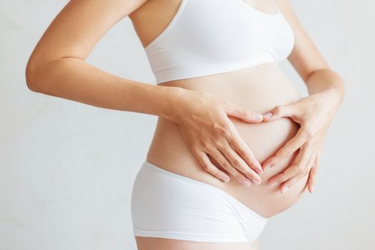 Pregnant woman in white underwear. Young woman expecting a baby. Future mother makes heart gesture - symbol of love.