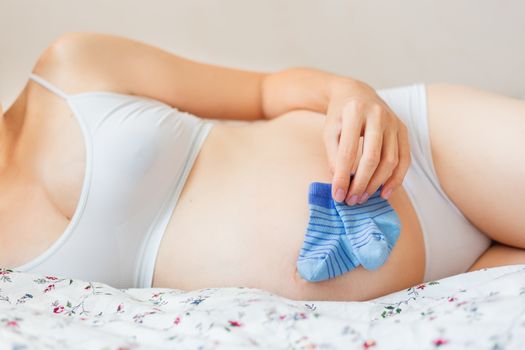 Pregnant woman in white underwear lying in bed with small baby's socks. Young woman expecting a baby boy.