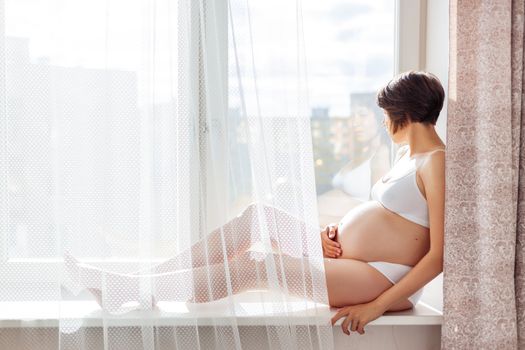 Pregnant woman in white underwear sitting on window sill. Young woman expecting a baby. Cozy happy background in sunny morning.