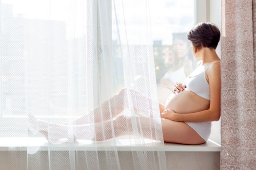 Pregnant woman in white underwear sitting on window sill. Young woman expecting a baby. Cozy happy background in sunny morning.