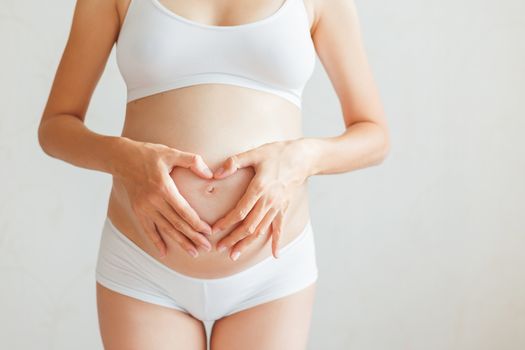 Pregnant woman in white underwear. Young woman expecting a baby. Future mother makes heart gesture - symbol of love.